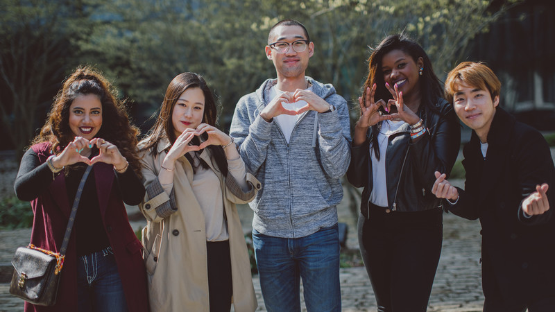 A group of international students stand in front of a building making heart shapes with their hands